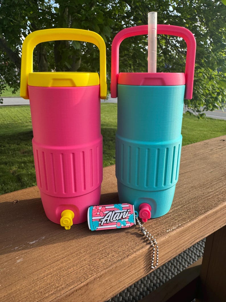 May include: Two insulated beverage coolers, one pink with a yellow handle and lid, the other teal with a pink handle and lid. A small Alani can-shaped charm is in front of the coolers. Both coolers have a spigot and a handle.