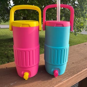 May include: Two colorful beverage coolers. One is pink with a yellow lid and handle, the other is teal with a pink lid and handle. Both have a spigot at the bottom and a handle at the top. A white straw is in the teal cooler.