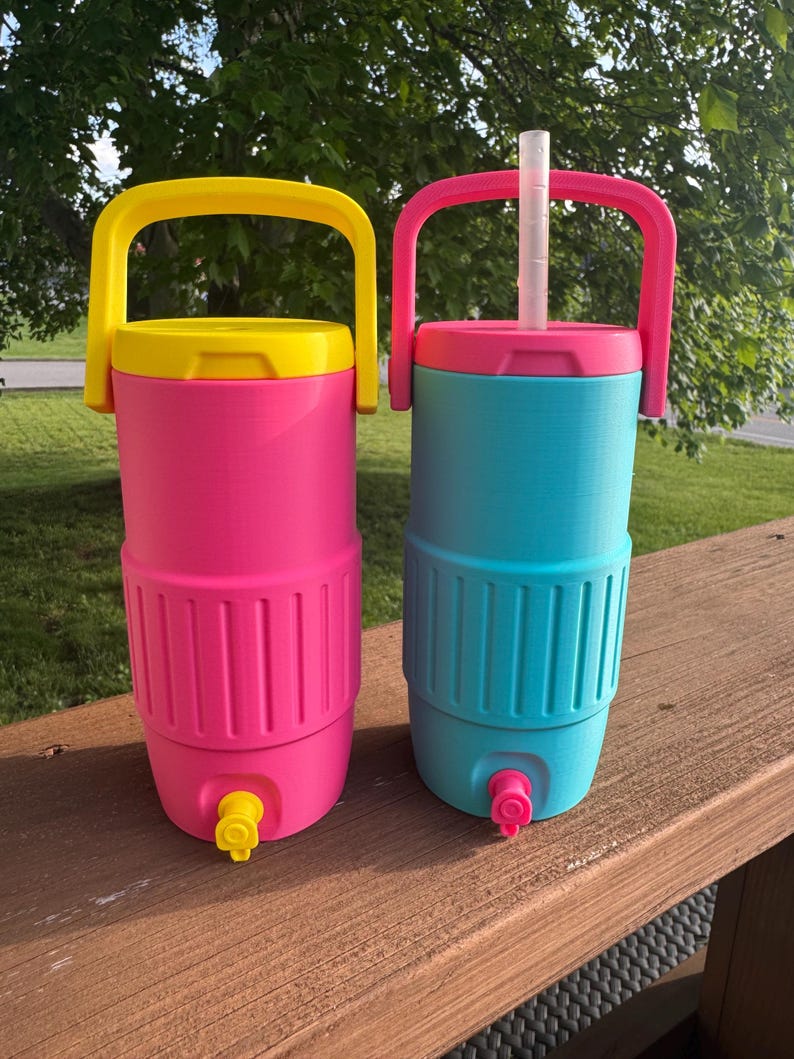 May include: Two colorful beverage coolers. One is pink with a yellow lid and handle, the other is teal with a pink lid and handle. Both have a spigot at the bottom and a handle at the top. A white straw is in the teal cooler.