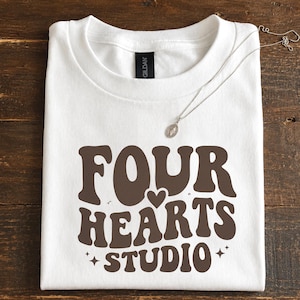 Könnte beinhalten: Ein weißes Kurzarm-T-Shirt mit dem braunen Schriftzug "FOUR HEARTS STUDIO" in einer wellenförmigen Schriftart, einem kleinen Herzsymbol zwischen "FOUR" und "HEARTS" sowie zwei kleinen vierzackigen Sternen neben dem Wort "STUDIO".