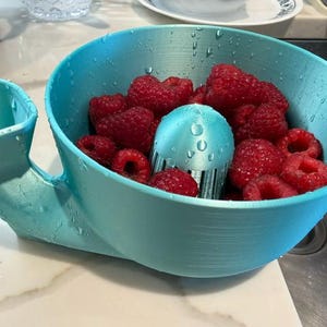 Peut inclure: Un saladier bleu clair imprimé en 3D avec une passoire intégrée. Le saladier est rempli de framboises rouges fraîches. La passoire est également bleu clair et a un design unique. Le saladier est posé sur un plan de travail blanc.