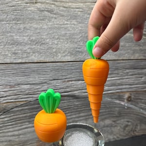May include: Orange and green carrot-shaped bottle stoppers. One stopper is held by a hand, the other sits on a clear, round base. The stoppers have a segmented orange body and green leafy tops. The background is a weathered wood.