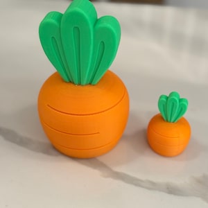May include: Two orange carrot-shaped ornaments with green tops. The larger carrot is in the foreground, and the smaller one is to the right. The carrots have horizontal lines around the body, and the green tops have a leaf-like design.