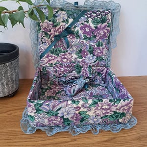 May include: A rectangular fabric-covered box featuring a floral pattern in shades of purple, green, and white. The interior and exterior are lined with this print, and the edges are trimmed with light blue lace. A dark green ribbon is attached inside.