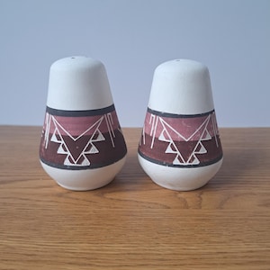 May include: A pair of matte white salt and pepper shakers with a tapered, rounded shape. Each shaker features a horizontal band around the center decorated with a dark red and black geometric pattern consisting of triangles and lines.