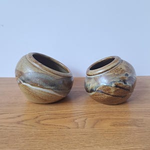 May include: Two round, asymmetrical ceramic bowls with a speckled, earthy glaze in shades of tan, brown, and grey. The bowls feature a slanted opening and a smooth, glossy finish, showcasing a marbled pattern across the exterior surface.