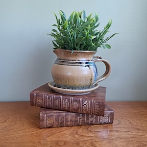May include: A ceramic pitcher with a tan and blue glaze sits on a small matching saucer, containing a green artificial plant. The pitcher rests on two stacked brown hardcover books titled Reader's Digest Books with gold lettering on the spines.