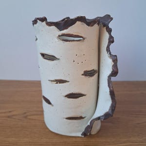 May include: A white ceramic vase with a birch bark design. The vase has a cylindrical shape with dark brown accents around the rim and edges. The vase has several horizontal oval cutouts and is approximately 18 cm tall.