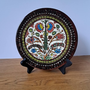 May include: A round ceramic decorative plate featuring a central floral motif with red, blue, and green petals on a cream background. The plate has a wide, dark brown border with a repeating white triangular pattern and sits on a black stand.