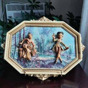 May include: An octagonal wall plaque featuring a cream-coloured ornate frame surrounding a three-dimensional scene. Two figures in period clothing are depicted against a painted landscape background, with one figure holding an umbrella and basket.