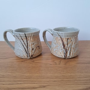 May include: Two ceramic mugs with a speckled light grey glaze and a textured tan lower section. Each mug features a hand-painted design of dark brown and white vertical lines resembling tall grass, with a curved handle on the side of each piece.