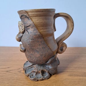 May include: A handcrafted ceramic mug featuring a sculpted face on the side with a prominent nose and eye detail. The mug has a two-tone brown glaze, a curved handle, and a base shaped like a large paw with visible toes and textured clay.