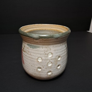 Peut inclure: Un pot à ail en céramique fait à la main, avec une finition blanc cassé mouchetée. Le récipient a une forme arrondie avec plusieurs trous circulaires et un bord vert foncé. Des accents floraux roses et verts subtils sont visibles.