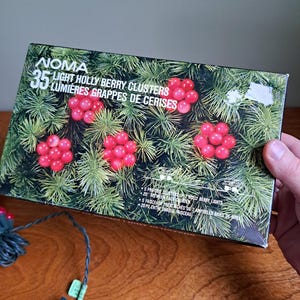 Peut inclure: Une boîte rectangulaire pour 35 lumières grappes de cerises NOMA avec un graphique de lumières rouges en forme de baies sur des aiguilles de pin vertes. Le texte indique 35 LUMIÈRES GRAPPES DE CERISES et 5 FAISCEAUX PRÉ-ATTACHÉS DE 7 AMPOULES BAIES DE BOIS.