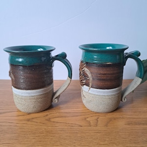 May include: Two handmade ceramic mugs. Each mug has a teal rim and handle, a brown middle section, a white stripe, and a tan base. One mug features a raised floral design. The mugs are on a wooden surface.