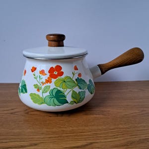 Peut inclure: Une casserole en métal émaillé blanc avec un couvercle assorti, ornée d'un motif floral composé de fleurs orange et de feuilles vertes. La casserole possède un manche en bois sur le côté et un petit bouton en bois au centre du couvercle.