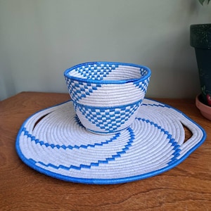 May include: A coiled basket and a matching circular tray featuring a white and bright blue geometric pattern. The basket has a tapered shape, while the flat tray includes two integrated handles, both constructed from tightly wound cord material.