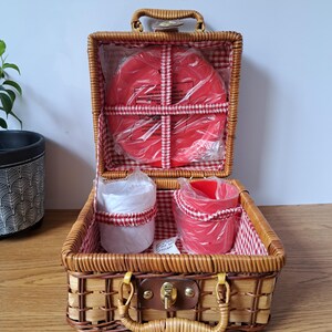 Peut inclure: Un panier de pique-nique en osier tressé avec un couvercle à charnière et un loquet en métal. L'intérieur est doublé d'un tissu à carreaux rouges et blancs. Il contient deux tasses en plastique, une blanche et une rouge, et une assiette rouge.