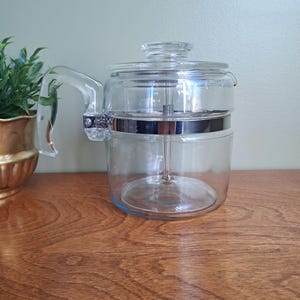 May include: A clear glass percolator coffee pot featuring a transparent lid, a sturdy glass handle, and a polished chrome metal band encircling the centre. The cylindrical vessel includes a central glass stem component visible inside the main chamber.