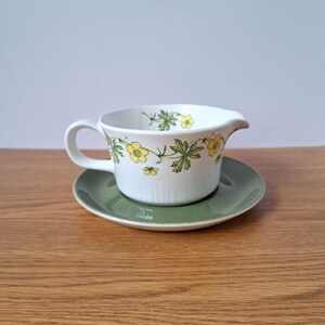 May include: A white ceramic creamer with a handle and a pouring spout, featuring a delicate botanical pattern of yellow flowers and green leaves around the rim. The creamer rests on a solid, muted olive green saucer with a smooth, circular edge.