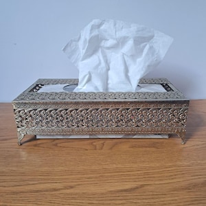 May include: A rectangular tissue box cover made of gold-toned metal with an intricate, openwork filigree pattern along the sides and top edges. The cover features four small, decorative feet and holds a white paper tissue extending from the centre.