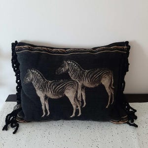 May include: Black rectangular decorative pillow featuring two zebras in a neutral color palette. The pillow has a decorative border and black fringe on the sides. The pillow is a home decor item.