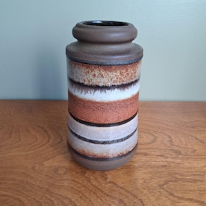 Peut inclure: Un vase cylindrique en céramique doté d'un bord épais brun foncé et d'un corps décoré de bandes horizontales dans des tons orange rouille, crème et brun foncé. La surface présente une finition texturée et mouchetée d'aspect mat.