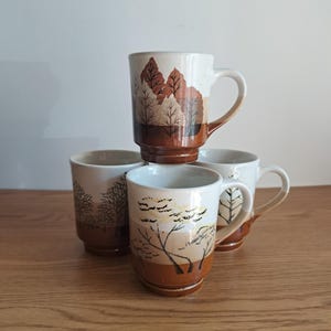 May include: A set of four ceramic mugs featuring a stacked design. Each mug has a white upper section and a brown base, decorated with brown and tan painted tree silhouettes. The mugs are arranged in a cluster with one mug positioned on top.