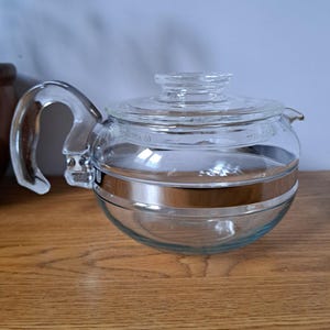 May include: A clear glass teapot featuring a rounded body, a matching glass lid, and a curved glass handle. A wide, metallic silver band encircles the center of the glass vessel, providing a distinct contrast to the transparent material of the pot.