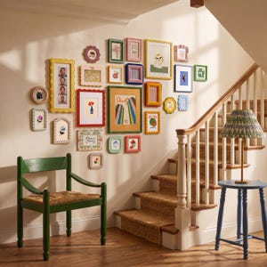 May include: A display of framed pictures and artwork in a variety of sizes and colours on a wall. A green chair with a woven seat is positioned below the wall art. A blue side table with a lamp and a staircase are also visible.