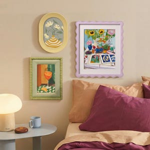 May include: Three framed wall art pieces featuring colorful illustrations of flowers, a drink, and a still life scene. The frames are oval yellow, rectangular green with a beaded edge, and rectangular lavender with a wavy scalloped border.