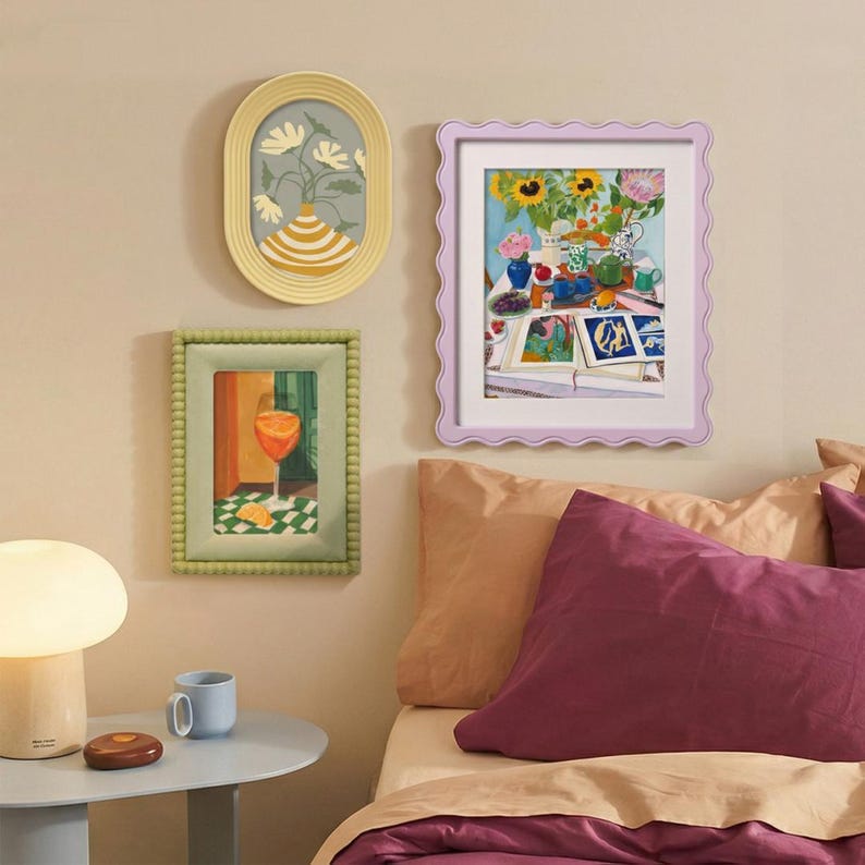 May include: Three framed wall art pieces featuring colorful illustrations of flowers, a drink, and a still life scene. The frames are oval yellow, rectangular green with a beaded edge, and rectangular lavender with a wavy scalloped border.