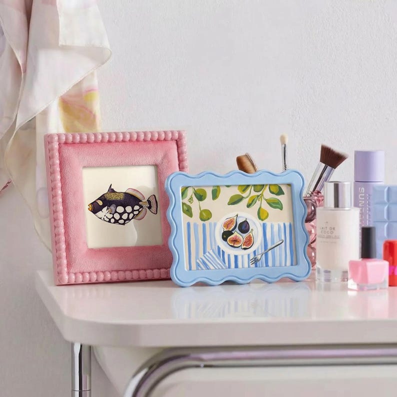 May include: A square pink beaded picture frame containing a fish illustration and a rectangular light blue wavy-edged picture frame featuring a painting of figs on a striped tablecloth, positioned next to various cosmetic bottles and brushes.