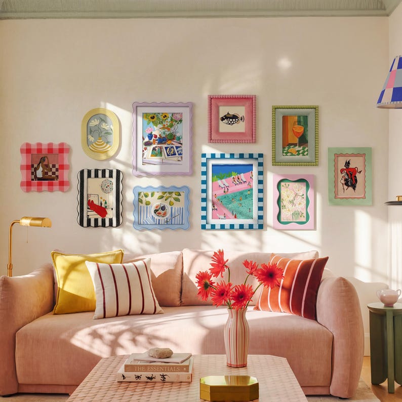 May include: A gallery wall featuring ten assorted picture frames in various shapes, colors, and patterns, including scalloped, striped, and beaded designs. The frames contain diverse art prints, arranged above a pink sofa with decorative pillows.