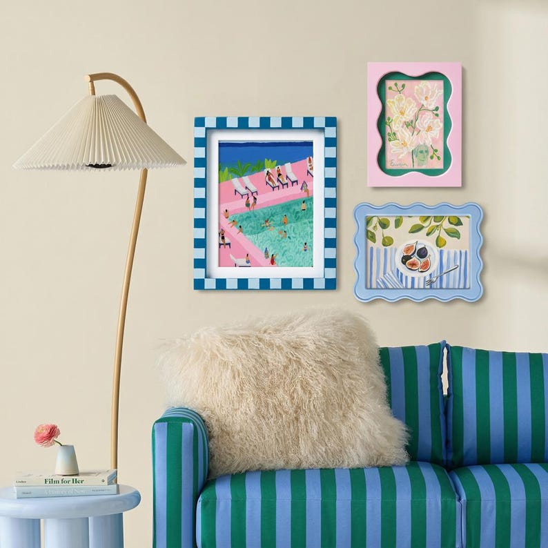 May include: A floor lamp with a pleated shade, a blue and white checkered frame containing a pool scene print, a pink wavy frame with a floral print, a light blue wavy frame with a fig print, and a green and blue striped sofa with a fluffy pillow.