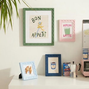 May include: A collection of framed art prints with a food and beverage theme. The prints include the phrases "Bon Appétit" and "Matcha Latte", along with images of a croissant and a coffee cup. The frames are in various colors, including green, pink, and blue.
