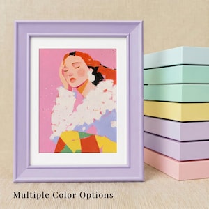 May include: A lavender picture frame containing an illustration of a person with orange hair, a white textured top, and a yellow and red patterned lower garment against a pink background, with a stack of frames in various pastel colors nearby.