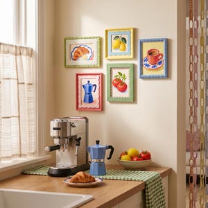May include: Five rectangular wall frames with colorful borders display illustrations of a croissant, lemons, a coffee cup, a blue moka pot, and tomatoes. Below, a stainless steel espresso machine sits next to a blue moka pot and a bowl of fruit.