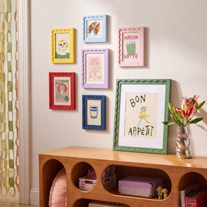 May include: A gallery wall arrangement of seven colorful wavy-edged picture frames containing various food and animal illustrations, including text reading Matcha Latte, Always Screamin, Cats & Pizza, and Bon Appétit, displayed above a wooden console.