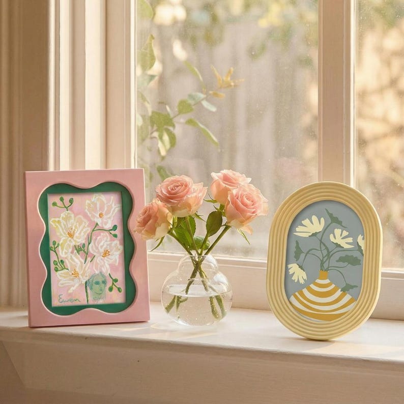 May include: A rectangular pink frame with a wavy inner edge containing a floral painting, a clear glass vase holding three light pink roses, and an oval yellow frame with a ribbed texture containing a print of a yellow and white striped vase.