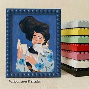 Puede incluir: Pintura enmarcada en azul que representa a una persona leyendo un libro y sosteniendo un gato. El marco tiene un borde de cuentas. La persona lleva una camisa floral. A la derecha hay una pila de bloques de construcción de colores. El texto en la parte inferior dice "Various sizes & shades."