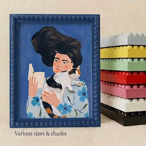 May include: A rectangular blue picture frame with a beaded border containing an illustration of a person with dark hair reading a book while holding a calico cat. A stack of identical frames in yellow, green, red, pink, white, and black sits nearby.
