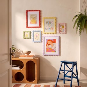 May include: A collection of six colourful, scalloped-edge picture frames arranged on a wall. The frames are red, yellow, light blue, pink, and lavender, containing various illustrations including text that reads NEVER STOP PLAYING.