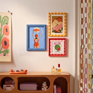 May include: Three framed wall art prints: a blue beaded frame with a swimmer holding a beach ball, a yellow checkered frame with a person sitting, and a red scalloped frame with a watermelon illustration labeled MELONE LA FRUTTA.