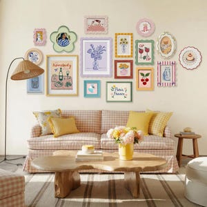 May include: A gallery wall featuring various framed art pieces in pastel colors. The frames have unique shapes and designs, including floral and scalloped edges. The artwork depicts illustrations of flowers, cats, and food. The wall is above a pink and white checkered sofa.