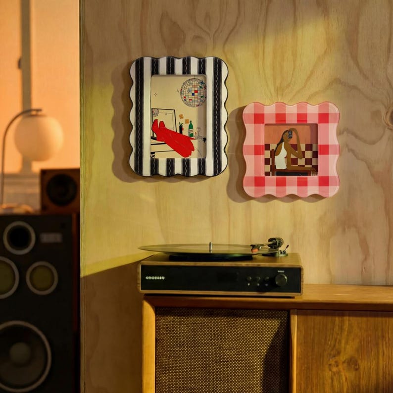 May include: Two wavy-edged picture frames mounted on a wall. The left frame is rectangular with black and white vertical stripes, containing an illustration of a disco ball and red boots. The right frame is square with a pink and white gingham pattern.