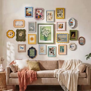 May include: A gallery wall of framed artwork in diverse shapes and sizes, mounted above a brown sofa. The frames display a range of colours and designs, including floral patterns, landscapes, and animal illustrations. The sofa is decorated with cushions and a blanket.