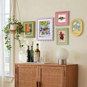 May include: A wooden cabinet with cane doors supports a white globe lamp, glass tumblers, and four bottles. Above, a hanging plant and five wall frames display various colorful illustrations, including a fish, a devil, and floral arrangements.