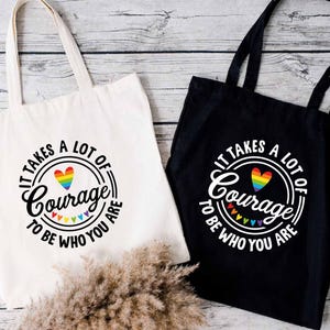 May include: Two tote bags, one white and one black, with the text "IT TAKES A LOT OF Courage TO BE WHO YOU ARE" in a circular design. The design includes a rainbow heart and rainbow triangles.