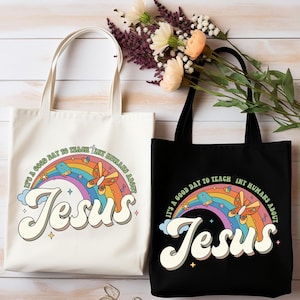 May include: Two tote bags, one cream and one black, with the text "It's a good day to teach tiny humans about Jesus" above the word "Jesus" in a retro font. A rainbow, butterflies, and clouds are also featured in the design.